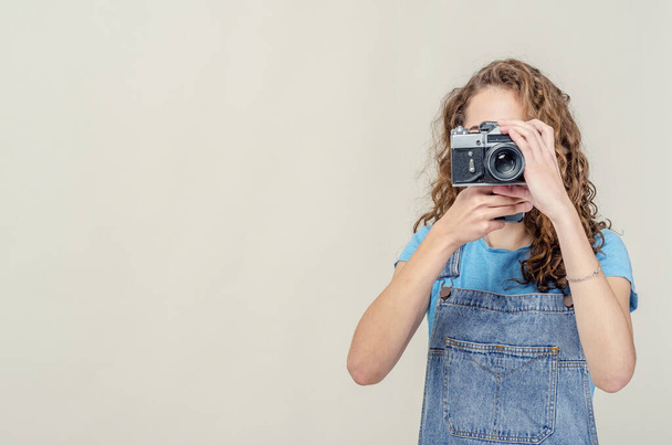 Curly brunette girl in denim overalls is holding a vintage film camera in her hands. Takes a picture - Fotoğraf, Görsel