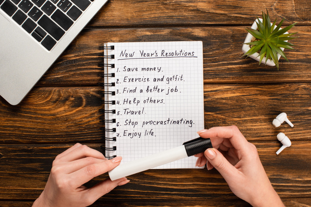 cropped view of businesswoman holding felt-tip pen near notebook with list of new years resolutions near laptop, potted plant and wireless earphones on wooden desk - Photo, Image