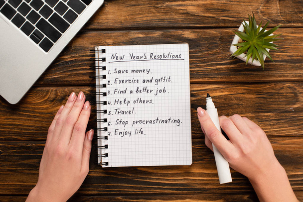 cropped view of businesswoman holding felt-tip pen near notebook with list of new years resolutions near laptop and potted plant on wooden desk - Photo, Image