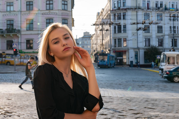 Chernivtsi, Ukrajina, 25.08.2019: Módní mladá Evropanka v černých šatech, stojící mezi starým centrem města a dívající se do kamery - Fotografie, Obrázek