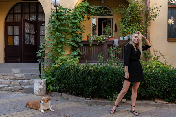 Menina loira sorridente bonita em vestido preto fica perto de um pequeno cão bonito, varanda velha decorada com vasos verdes no fundo
 - Foto, Imagem