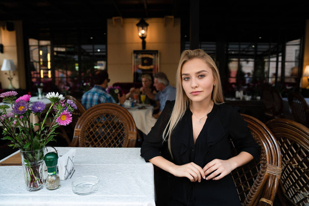 Chernivtsi, Ucrânia, 25.08.2019: Menina loira bonita elegante em vestido preto sentado em uma mesa em um café acolhedor rua decorada com flores
 - Foto, Imagem