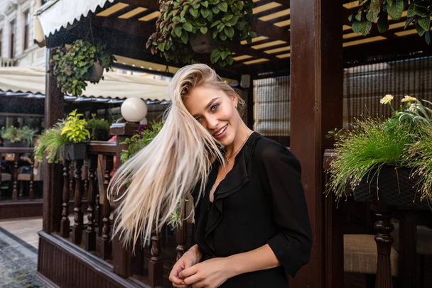 Beautiful young blonde woman standing with soaring hair near the summer terrace decorated with green flowerpods - Photo, image