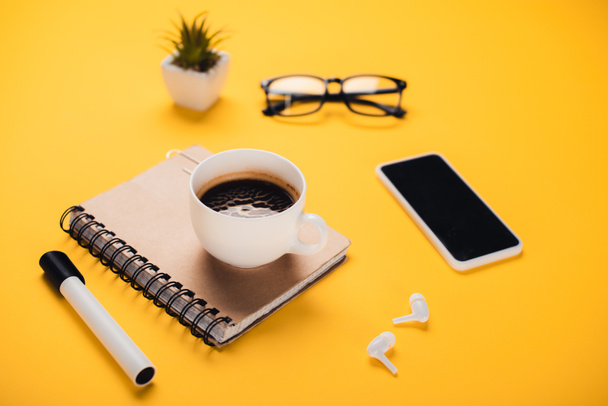 coffee cup on notebook near smartphone, wireless earphones, glasses, potted plant and felt-tip pen on yellow desk - Photo, Image