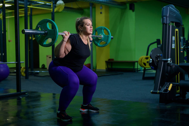 Overweight woman crouches with a barbell in the gym. A fat girl does exercises on the buttocks. Goes in for sports for weight loss. - 写真・画像