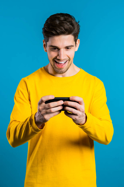 Handsome man playing game on smartphone on blue studio wall. Using modern technology - apps, social networks. - Φωτογραφία, εικόνα
