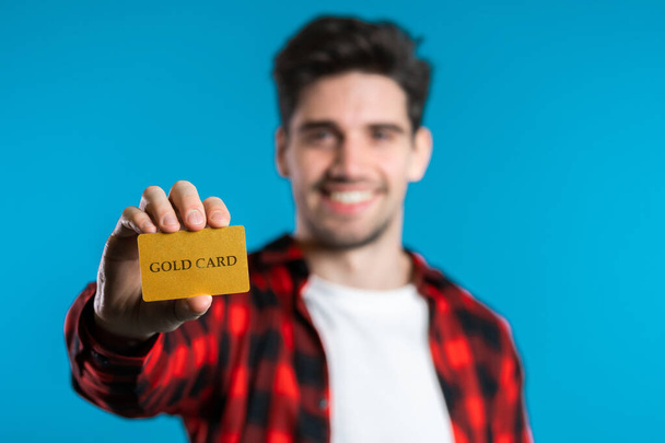 Successful man in plaid shirt showing unlimited gold credit card on blue studio background. Student, money concept - Foto, afbeelding