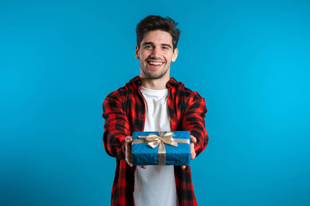 Handsome man holding christmas gift and gives it by hands it to camera. He is happy, smiling. Guy on blue background. Positive holiday shot - Photo, image