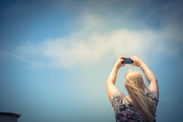 Vista de pesquisa da jovem senhora caucasiana tirando fotos usando seu telefone inteligente. Menina loira de baixo ângulo com telefone celular sob céu azul nuvem de verão
 - Foto, Imagem
