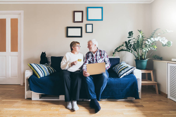 Пожилая пара делает покупки онлайн на диване дома, глядя друг на друга и улыбаясь
 - Фото, изображение