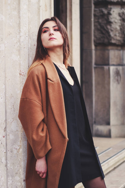 Model in zwarte jurk en lange bruine jas. Een moersleutel. Stijl. Mode - Foto, afbeelding