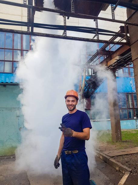 Portrait of a worker in a factory. Steam under pressure in the background. in a helmet and gloves examines the territory of the genus Palomak. elimination of defects in production. professionalism in  - Photo, Image