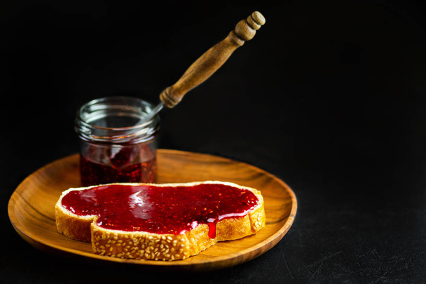 Bread with jam on a black background. Raspberry jam on white bread in a wooden plate. Breakfast toast with raspberry jam. Vegetarian breakfast. Vegan snack - Photo, image