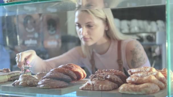 Gyönyörű fiatal női eladó, aki a cukrászdában dolgozik. Friss péksütemények közelsége vitrinen - Felvétel, videó