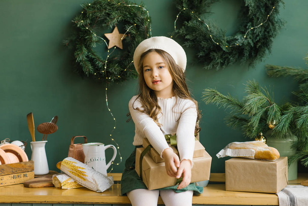 Een stijlvol meisje in een witte baret zit in een stijlvolle keuken ingericht voor Kerstmis en Nieuwjaar in smaragd of groen. Kerstkransen aan de muur. Meisje met een kerstcadeau - Foto, afbeelding