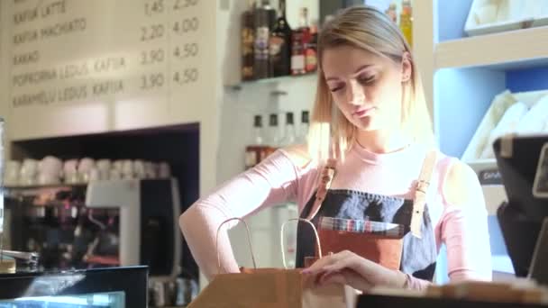 Όμορφη νεαρή κοπέλα πωλητής συσκευάζει κέικ στέκεται στον πάγκο στο ζαχαροπλαστείο - Πλάνα, βίντεο