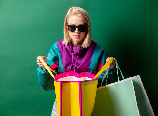 Blonde girl in 90s style jacket with shopping bags on green back - Φωτογραφία, εικόνα