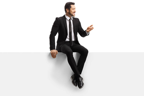 Handsome young man in a suit sitting on a blank panel and gestur - Photo, image