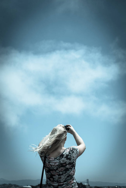 Vintage tone rear view of young Caucasian lady taking photo using her smart phone. Low angle blonde girl wears shoulder strap bag use cell phone under summer cloud blue sky - Photo, Image