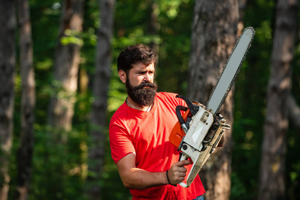 Firewood as a renewable energy source. Stylish young man posing like lumberjack. Firewood as a renewable energy source. Lumberjack with chainsaw on forest background. - Fotoğraf, Görsel