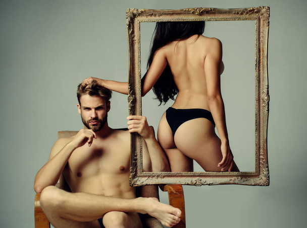 Bare hot girl with sexy ass standing in a frame. Handsome muscular bearded man holding frame with sexy hot woman. Abstract art. Sexy couple isolated at grey background. - Foto, Bild