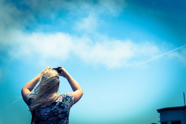 Rear view of young Caucasian lady taking photo using her smart phone. Low angle blonde girl wears shoulder strap bag use cell phone under summer cloud blue sky - 写真・画像