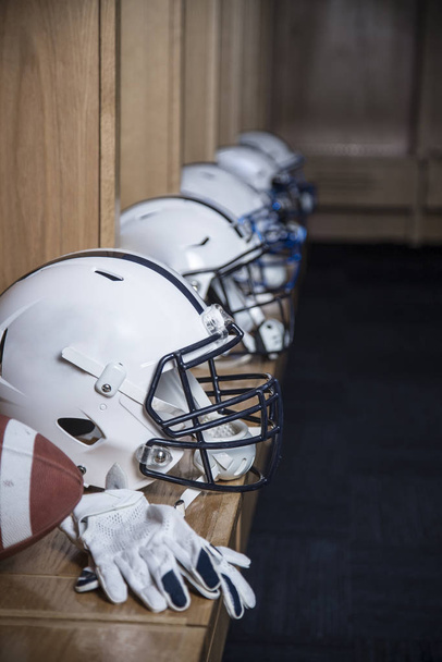 Nahaufnahme einer Reihe von American-Football-Helmen, die vor einem Fußballspiel in einer Umkleidekabine sitzen. Handschuhe und ein Fußball auch auf der Bank in der Umkleidekabine - Foto, Bild