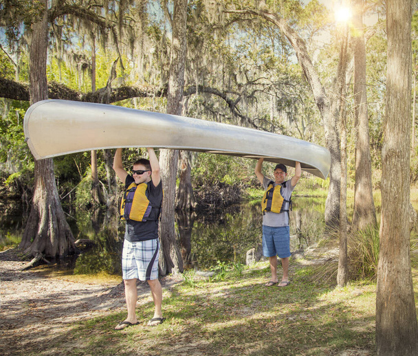 Twee avontuurlijke mannen klaar om te kanoën op een tropische rivier terwijl ze hun kano lachend het water in dragen en klaar voor een leuke tijd in de buitenlucht - Foto, afbeelding