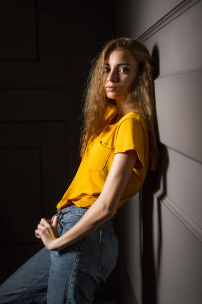 Beautiful young girl in a yellow T-shirt and jeans stands against a wall in a dark room - Photo, image