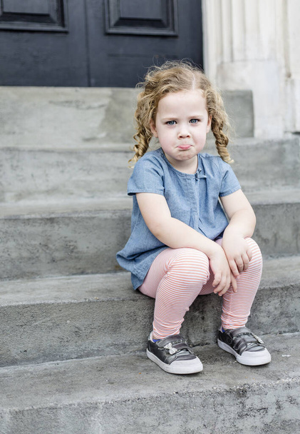 Emotionales Porträt eines traurigen, unglücklichen kleinen Mädchens mit blonden lockigen Haaren, das auf den vorderen Stufen ihres Hauses in einer städtischen Umgebung sitzt. Netter Ausdruck und entzückendes Gesicht. Kinderverhaltenskonzept Foto - Foto, Bild