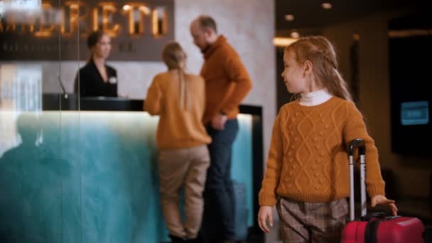 Una familia registrándose en el hotel de lujo - una niña pequeña parada con una maleta y esperando a sus padres - volviendo la cabeza para ver a sus padres
 - Imágenes, Vídeo