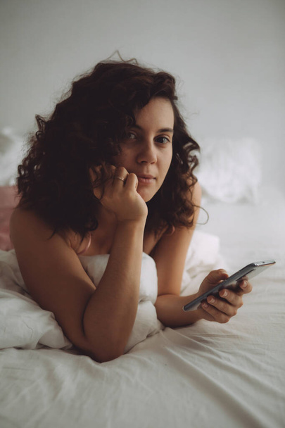 young pretty woman with curly hair in bed with phone - Φωτογραφία, εικόνα