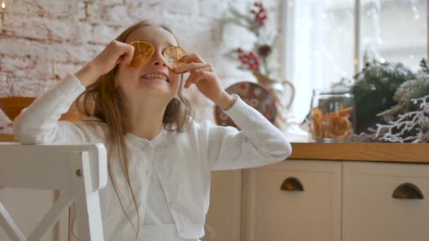Hymyilevä pieni vaalea tyttö grimacing ja peittää silmänsä kuivattuja appelsiininviipaleita kotona parvi tyyli ja joulukoristeen taustalla, iloinen Xmas käsite
 - Materiaali, video