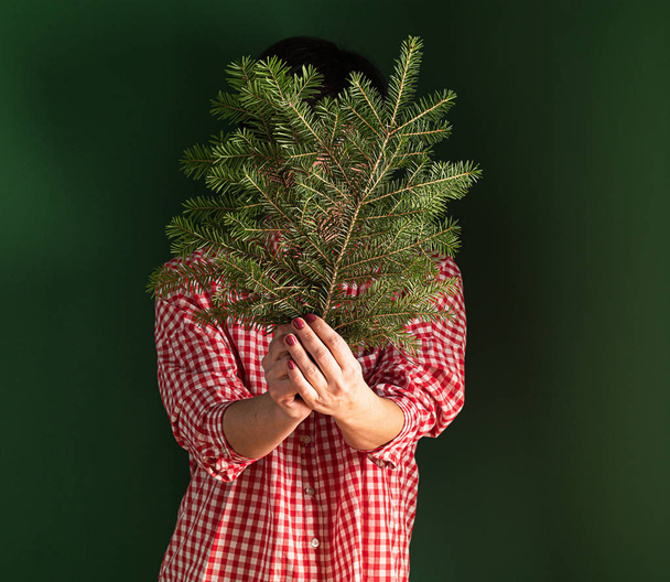 A woman red shirt is standing against a green wall, holding a Christmas tree branch in her hands and covering her face. - Foto, Imagen