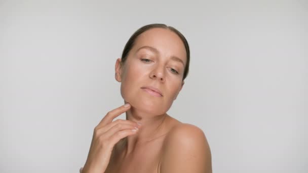 Close-up beauty portrait of young woman with smooth healthy skin, she gently touches her shoulders with her fingers on white background and smiles - Filmati, video