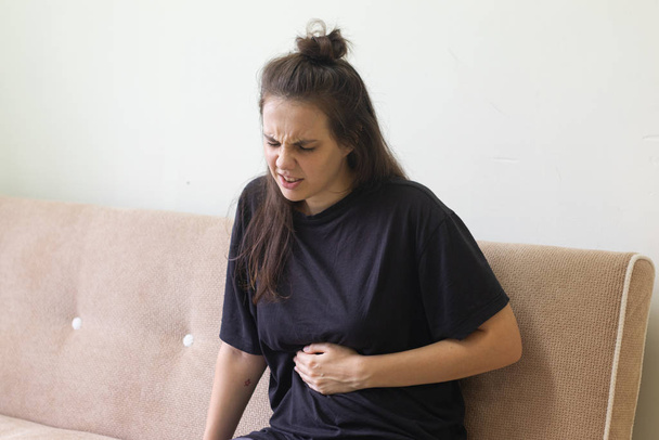 Молода жінка, яка страждає від болю в животі, сидить вдома на дивані
 - Фото, зображення