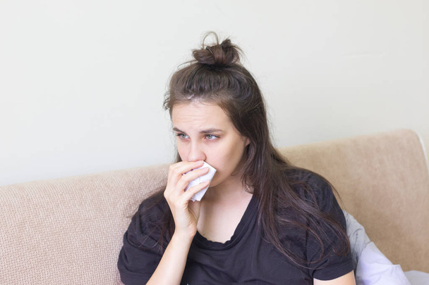 Άρρωστη νεαρή γυναίκα κάθεται στο σπίτι με μια καταρρέουσα μύτη. - Φωτογραφία, εικόνα