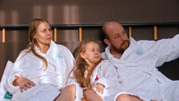 Μια ευτυχισμένη οικογένεια με λευκά μπουρνούζια που κάθεται στο κρεβάτι και μιλάει ο ένας στον άλλο. - Πλάνα, βίντεο