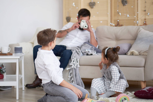 Ο μπαμπάς και τα παιδιά διασκεδάζουν μαζί στο σπίτι, ο μπαμπάς κάθεται στον καναπέ και φωτογραφίζει τα παιδιά του. Ο γιος και η κόρη παίζουν στο πάτωμα. - Φωτογραφία, εικόνα