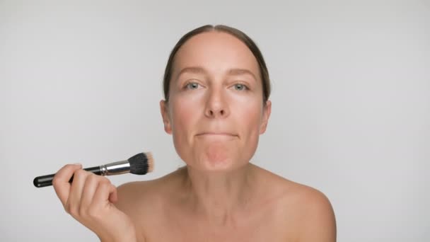 Close-up beauty portrait of young woman with smooth healthy skin, she gently strokes the skin with a makeup brush on white background - Materiał filmowy, wideo