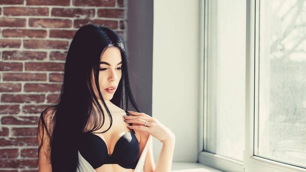 The woman of admire. Sexi woman with makeup and sensual look. Sexy woman in erotic underwear standing at window. Pretty woman with long brunette hair - 写真・画像