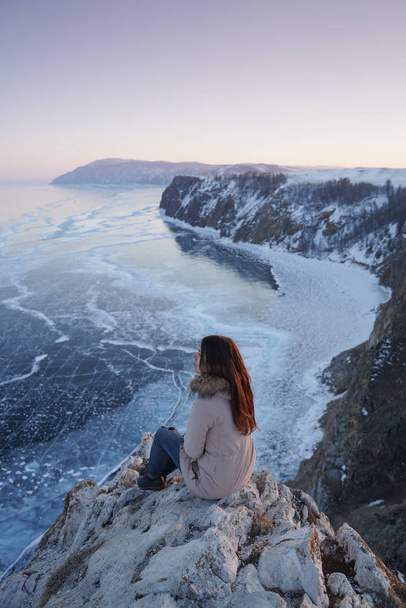 Lake Baikal in winter. Woman sitting on a edge of cliff and looking at frozen Baikal lake. - Photo, Image