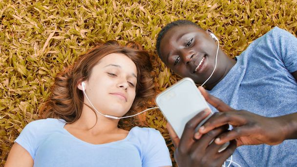 Multi ethnic couple of teenagers lying on the grass hearing music. Happiness and lightheartedness concept. - Photo, Image