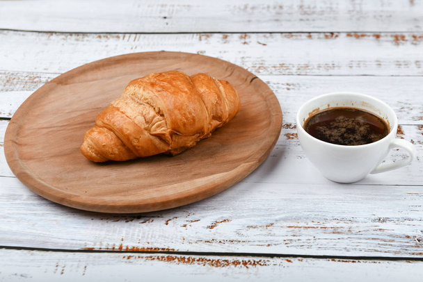 Croissant und Tasse Kaffee auf einem weißen Holztisch. Frisches französisches Croissant. auf einem hölzernen Hintergrund. Blick von oben. Morgens Frühstück mit einem Croissant. Französisches Frühstück zu Hause zubereitet. - Foto, Bild