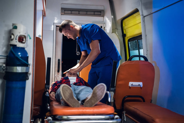 Man in medisch uniform zet zuurstofmasker op zinloze vrouw liggend op brancard in de ambulance auto - Foto, afbeelding