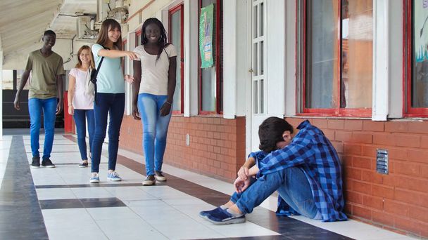Young caucasian sitting alone with sad feeling at school. Child in depression abandoned in a corridor and leaning against brick wall. Bullying, discrimination and racism concept. - Foto, afbeelding