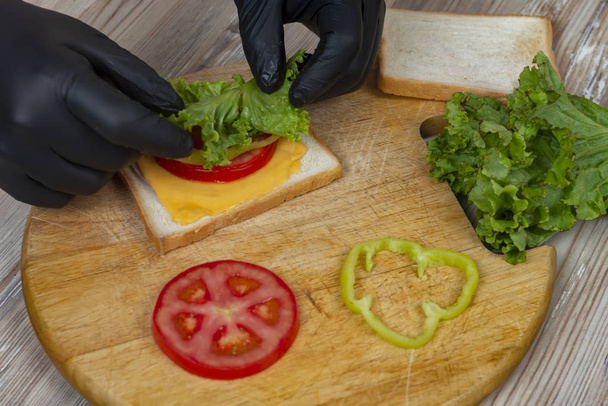 άνδρας με μαύρα γάντια προετοιμάσει σάντουιτς σε ξύλινο γραφείο - Φωτογραφία, εικόνα