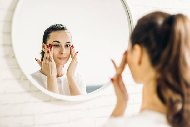Όμορφη γυναίκα που βάζει καλλυντικά στο πρόσωπό της. Γυναίκα στο μπάνιο εφαρμογή κρέμα στο πρόσωπο. - Φωτογραφία, εικόνα