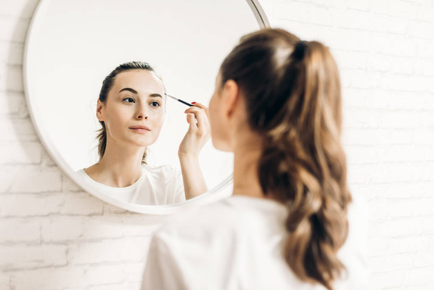 Portrait of beautiful smiling woman looking in mirror and applying mascara. - Фото, зображення