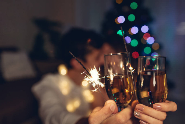 Christmas and New year party concept. Couple in love burning sparklers by illuminated Christmas tree with champagne. - Fotó, kép